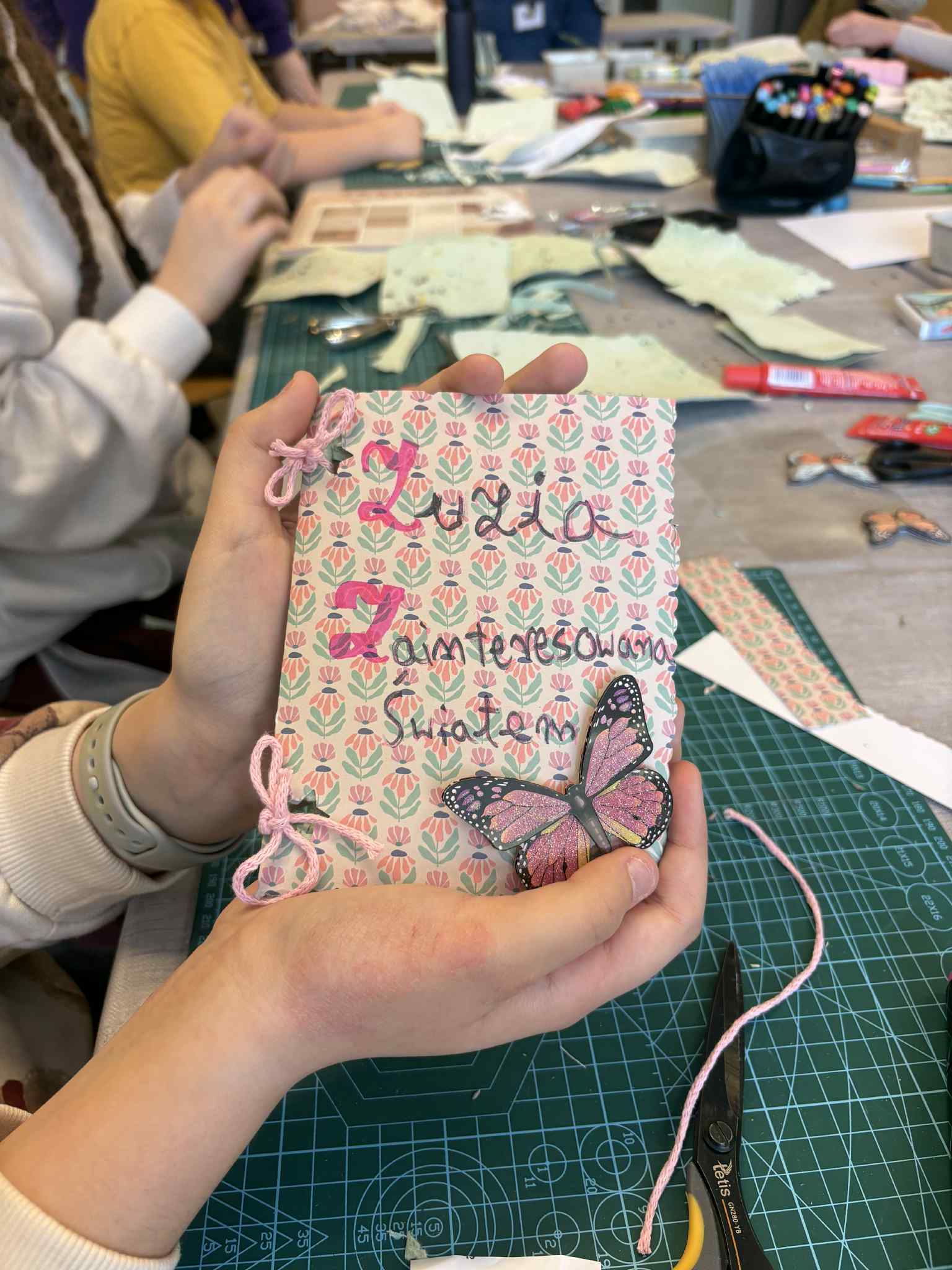 Dłonie trzymają ręcznie wykonaną książeczkę z motylem i napisem na okładce.