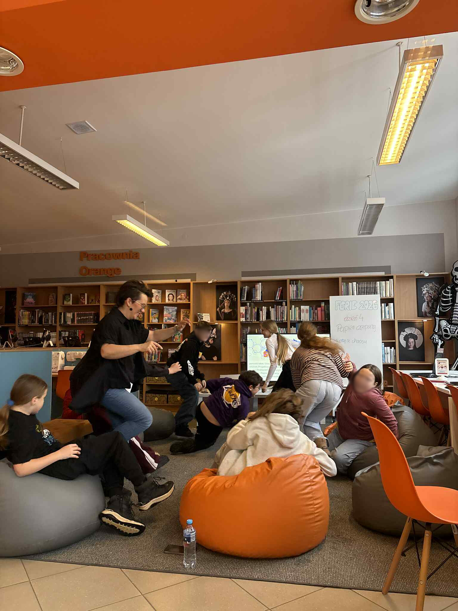 Grupa dzieci i prowadząca bawią się i poruszają na pufach w bibliotece.