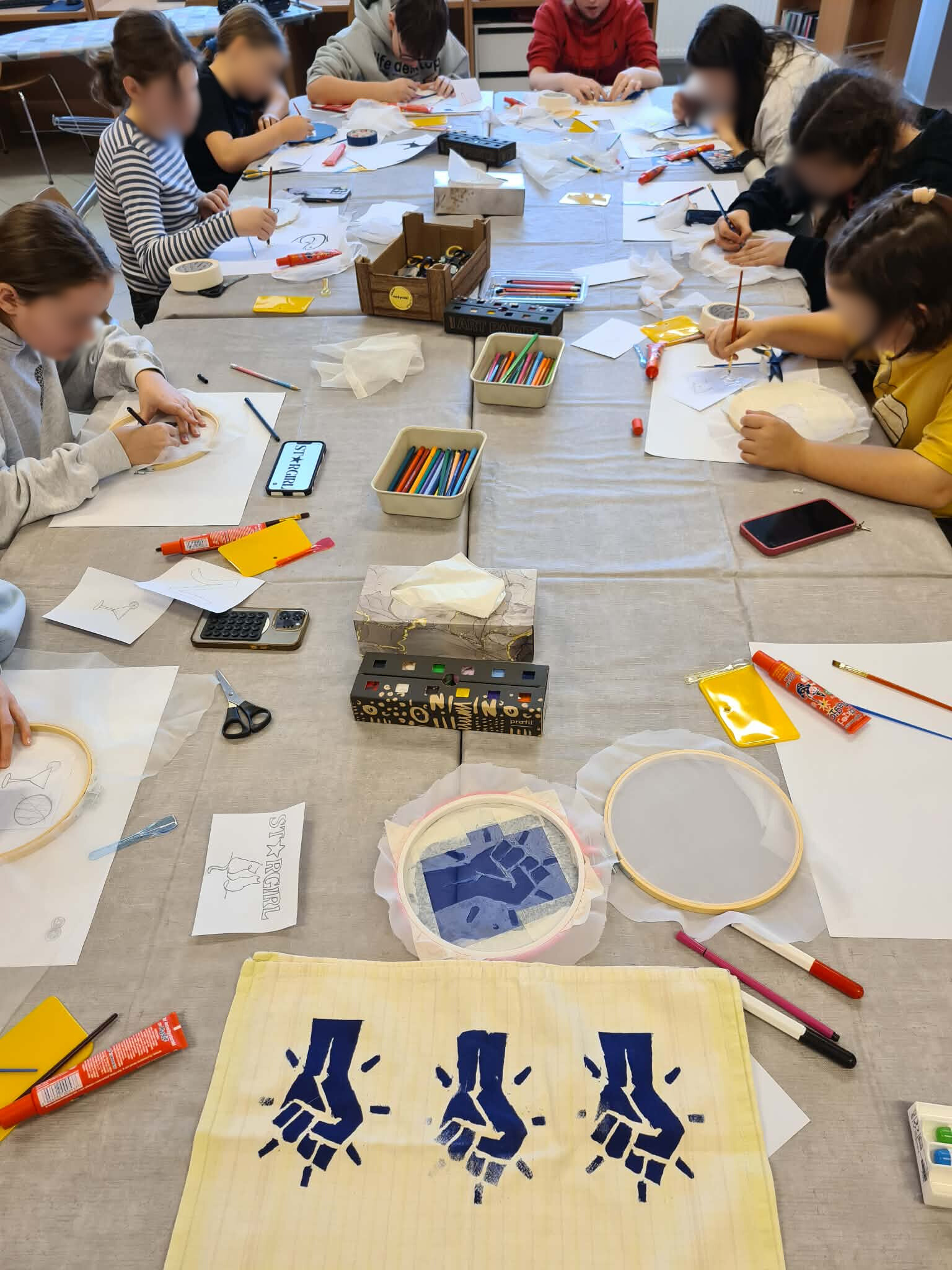 Dzieci przy dużym stole tworzą prace plastyczne; na pierwszym planie materiałowa torba z trzema granatowymi nadrukami dłoni, wokół przybory plastyczne.