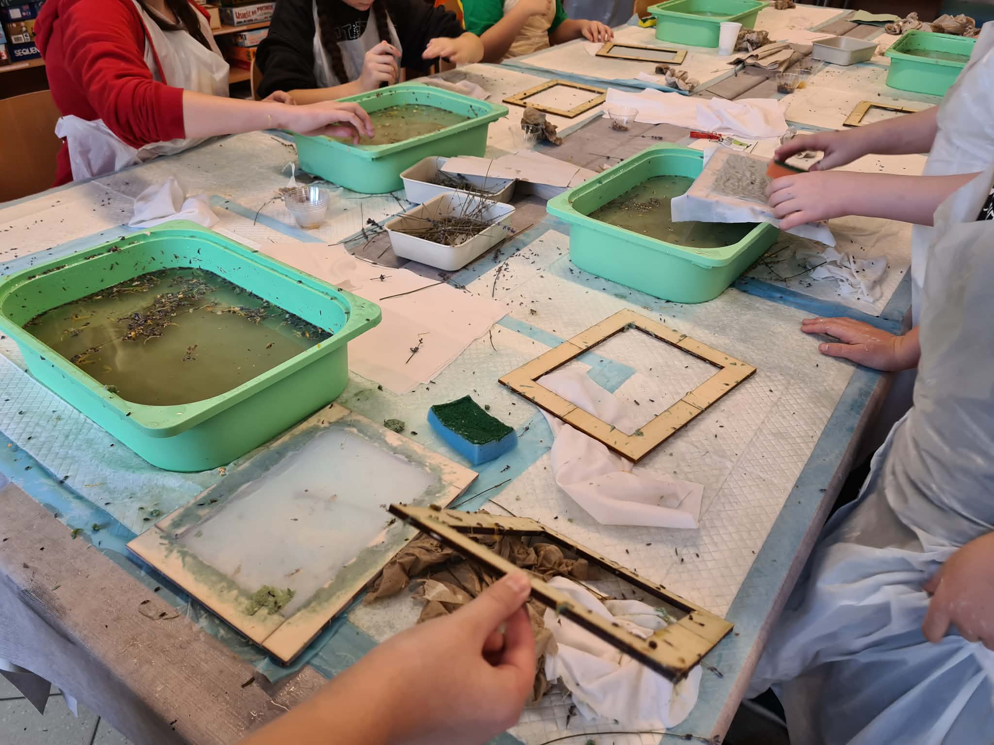 Dzieci pracują przy stole, czerpiąc papier w zielonych miskach z masą papierową.