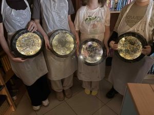 Dzieci stoją obok siebie i trzymają okrągłe, błyszczące prace plastyczne przypominające Księżyc.