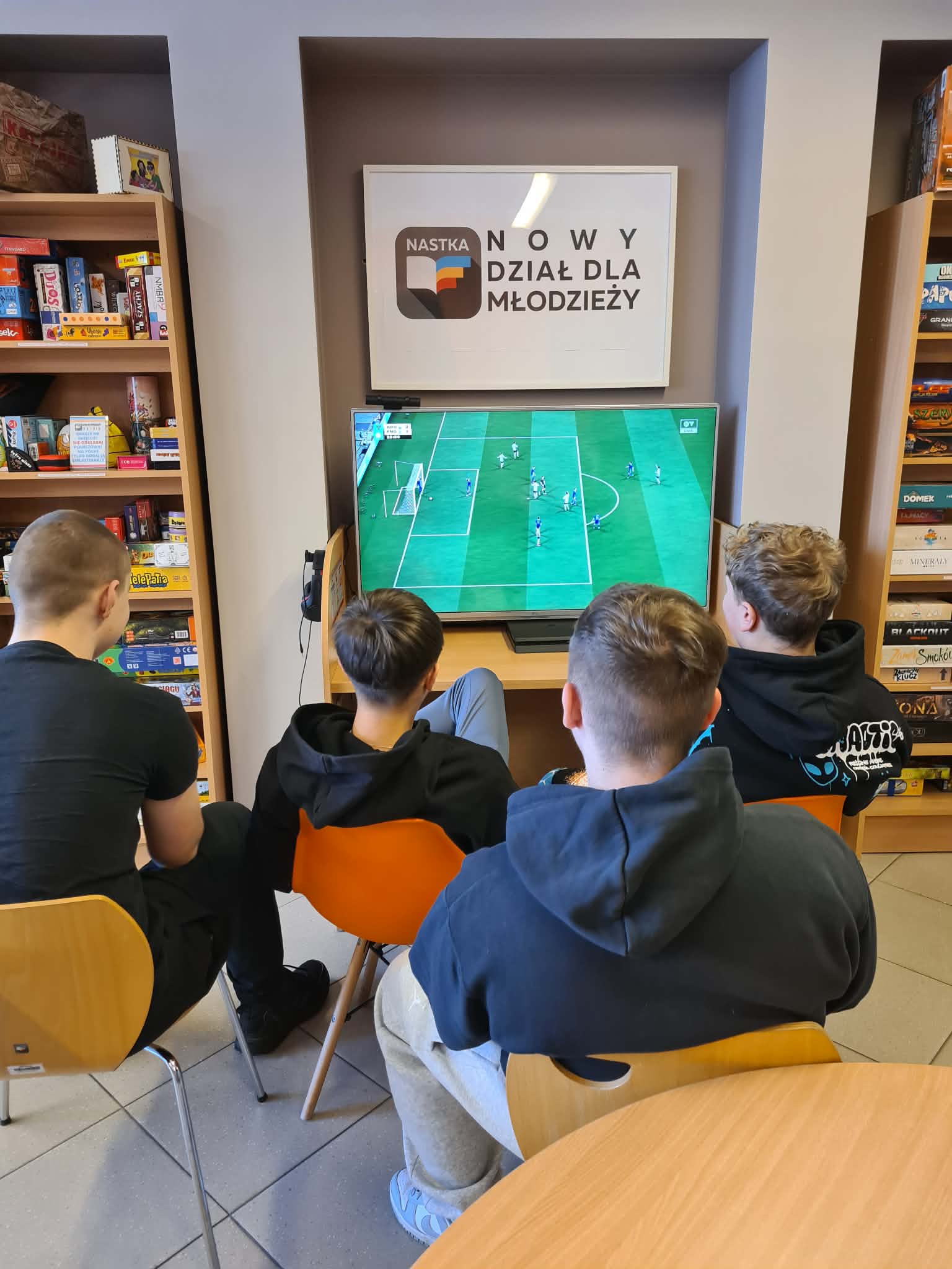 Kilku chłopców siedzi przed dużym ekranem i gra w grę piłkarską. Nad monitorem widoczny napis „Nowy Dział dla Młodzieży NASTKA”.