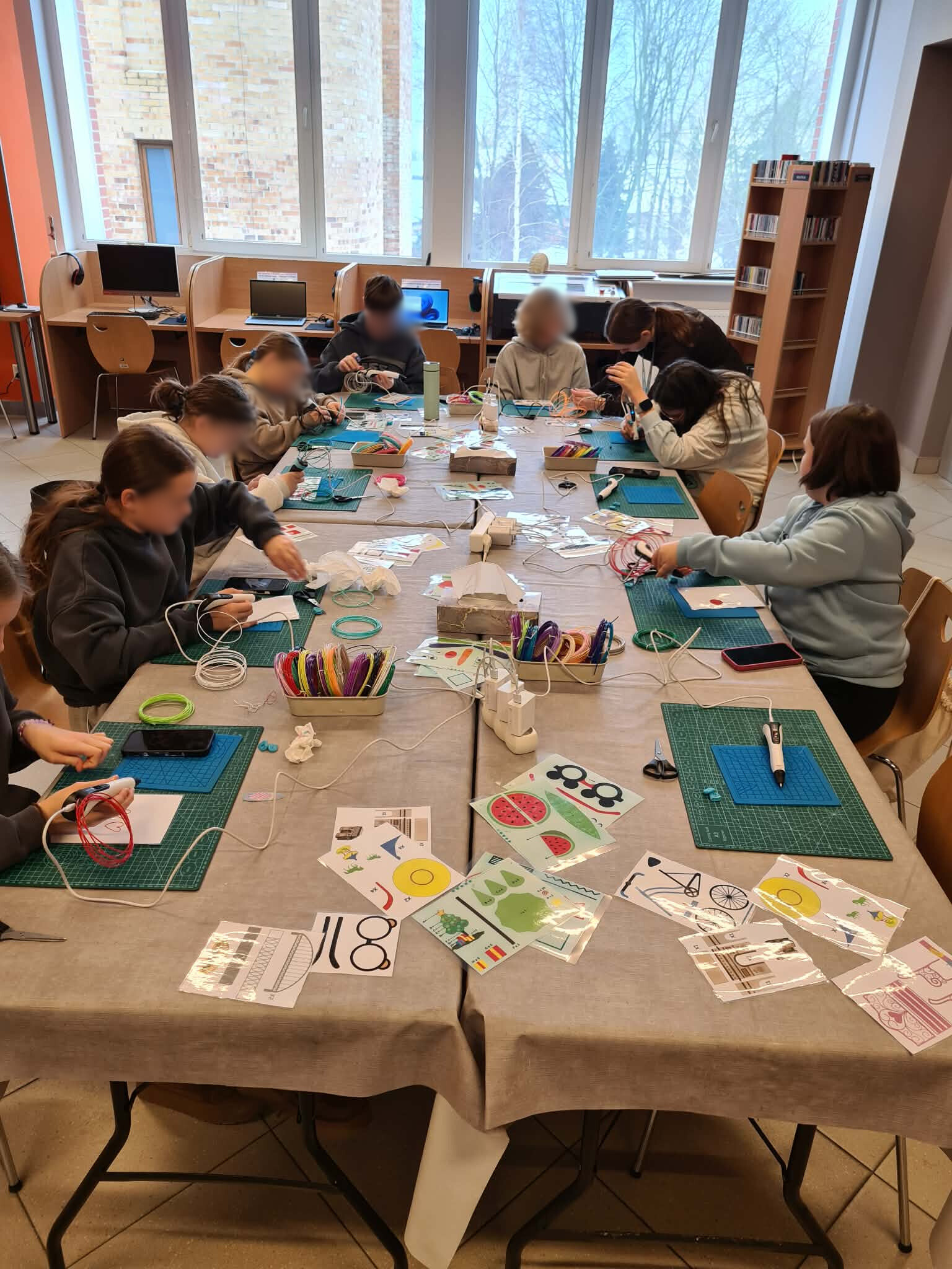 Grupa młodzieży pracuje przy stołach warsztatowych z długopisami 3D i materiałami.