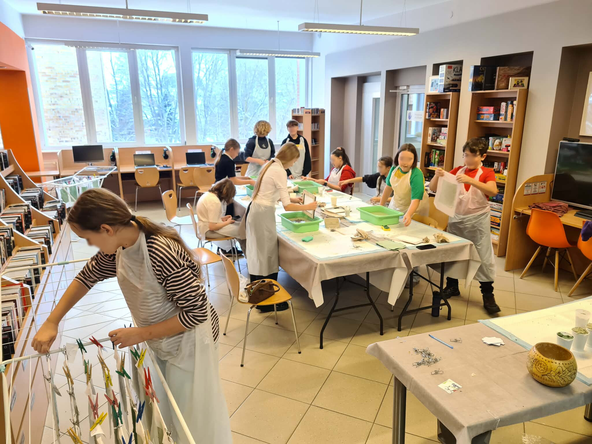 Uczestnicy warsztatów w fartuchach tworzą papier czerpany w przestrzeni biblioteki, część suszy arkusze.
