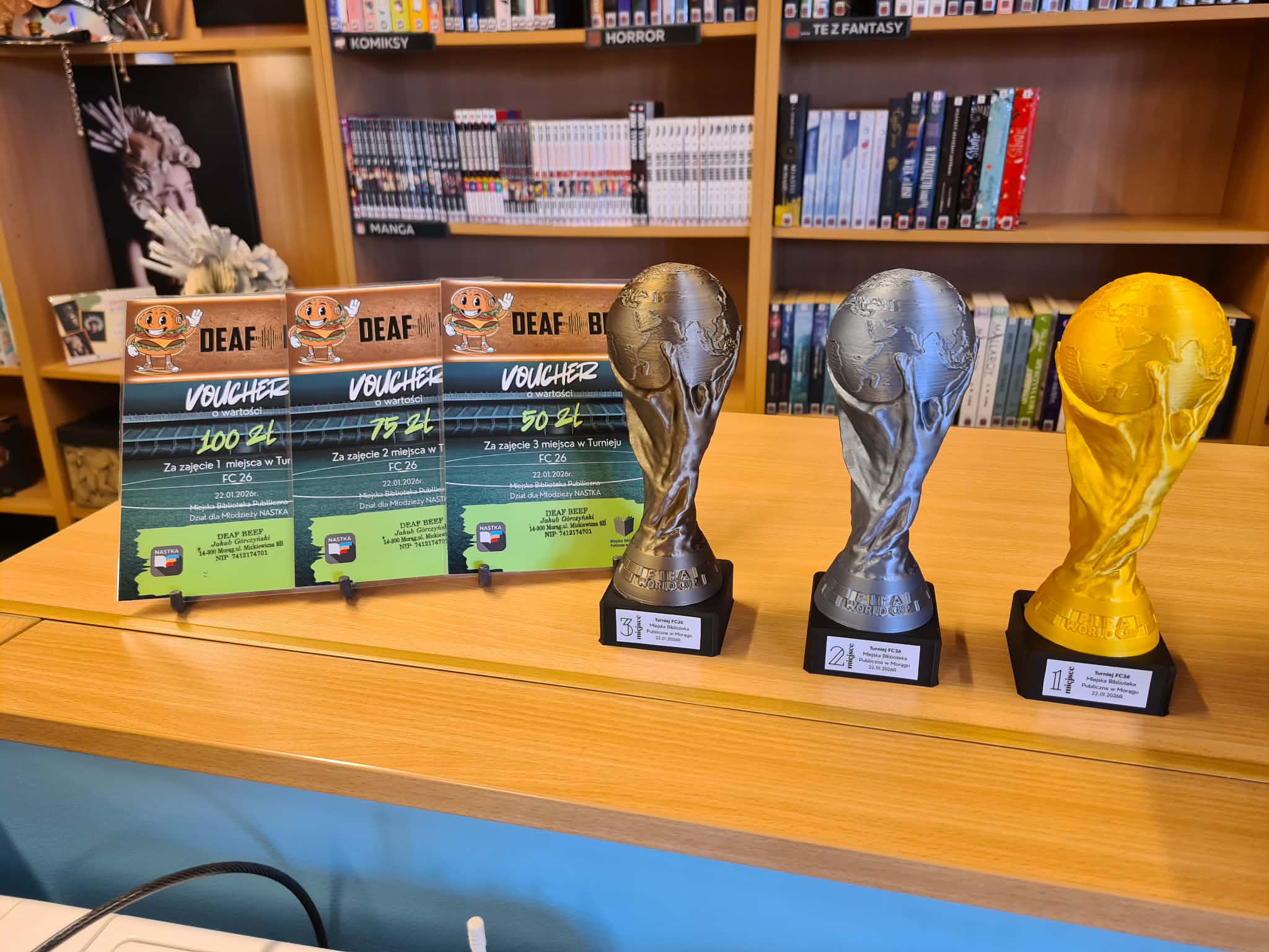 Na ladzie w bibliotece ustawione są nagrody z turnieju FC 26: trzy vouchery o różnych wartościach oraz trzy puchary w kształcie piłkarskiego trofeum w kolorach złotym i srebrnym. W tle regały z książkami.