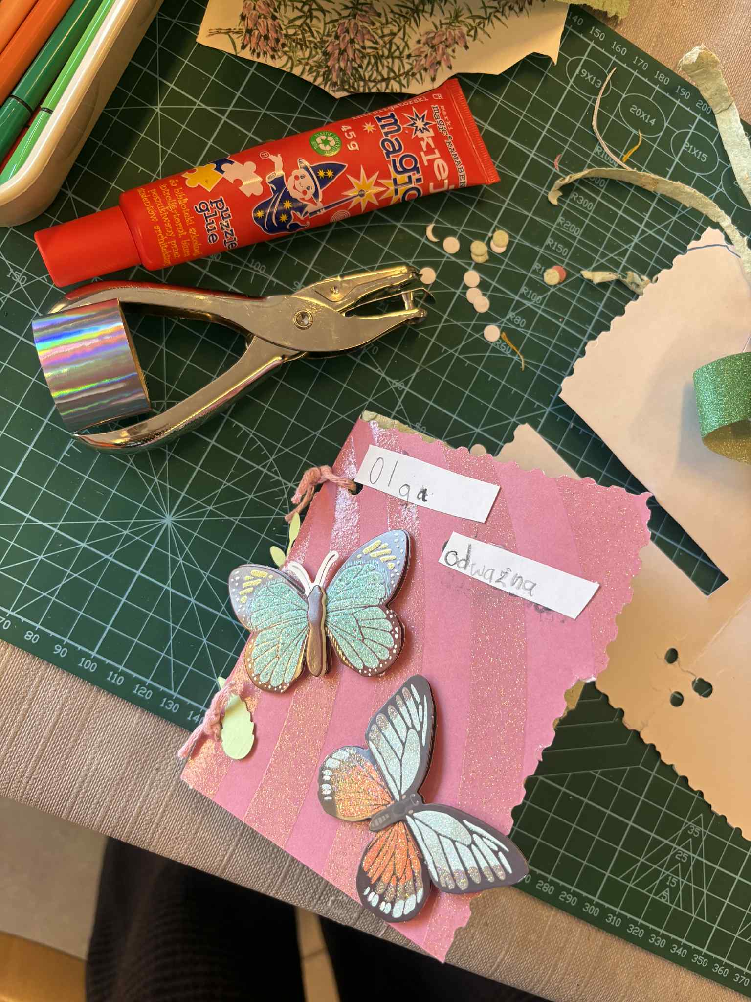 Ozdobiona okładka książeczki z papieru czerpanego z motylami i podpisami, obok narzędzia plastyczne.
