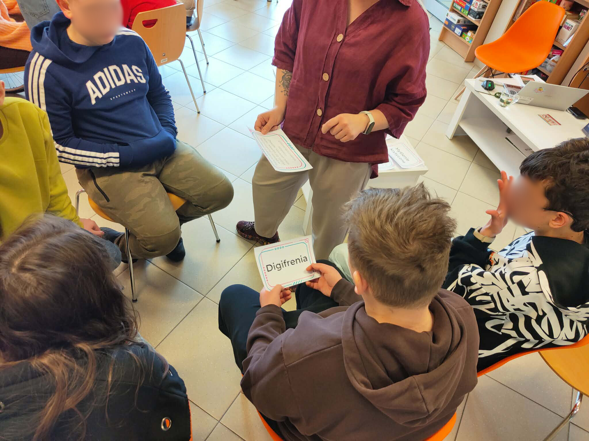 Młodzież siedzi w kręgu podczas ćwiczenia, prowadząca rozdaje karty z hasłami.