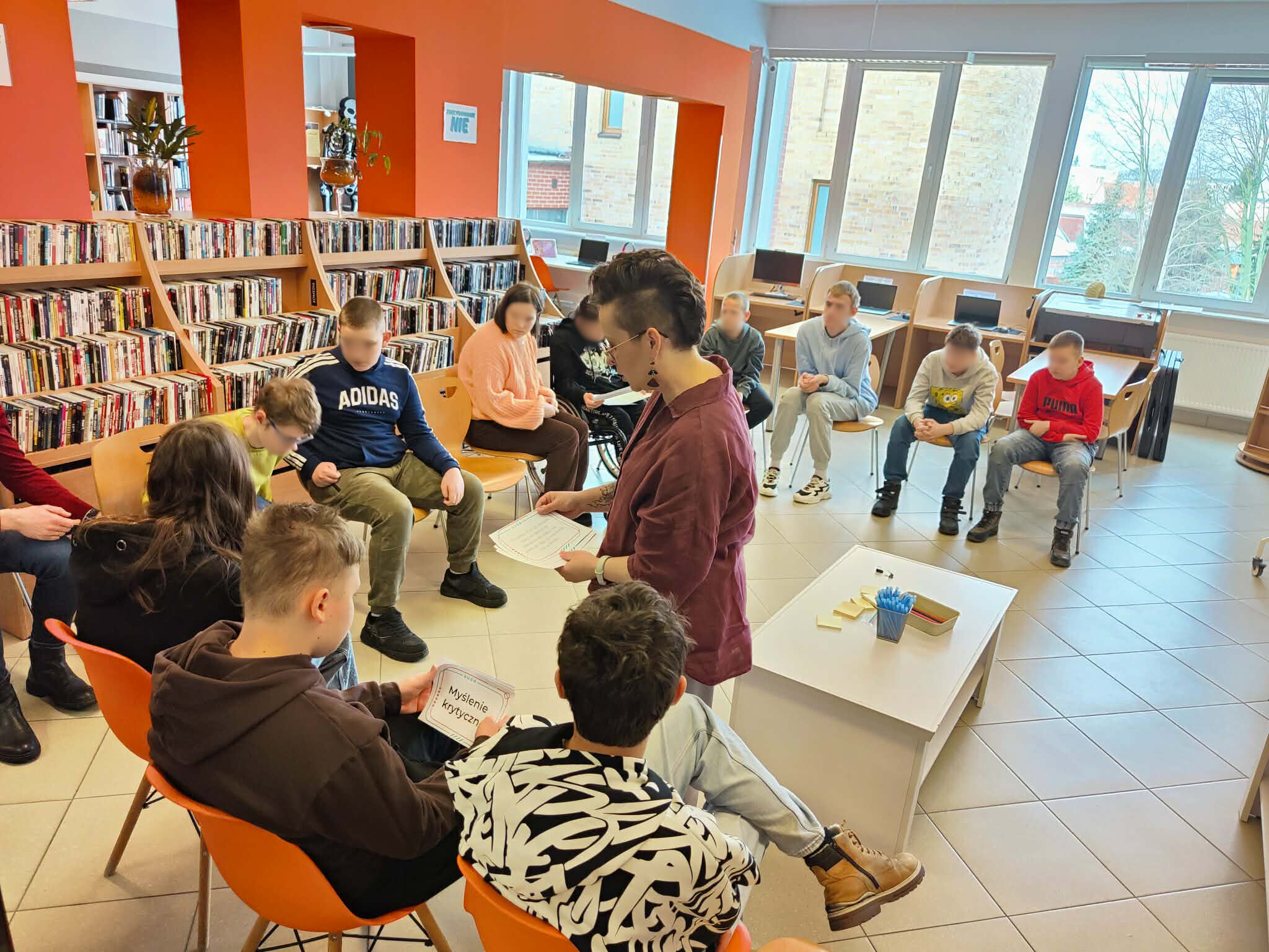Zajęcia w dziale dla młodzieży – grupa siedzi w kręgu, prowadząca rozdaje materiały edukacyjne.