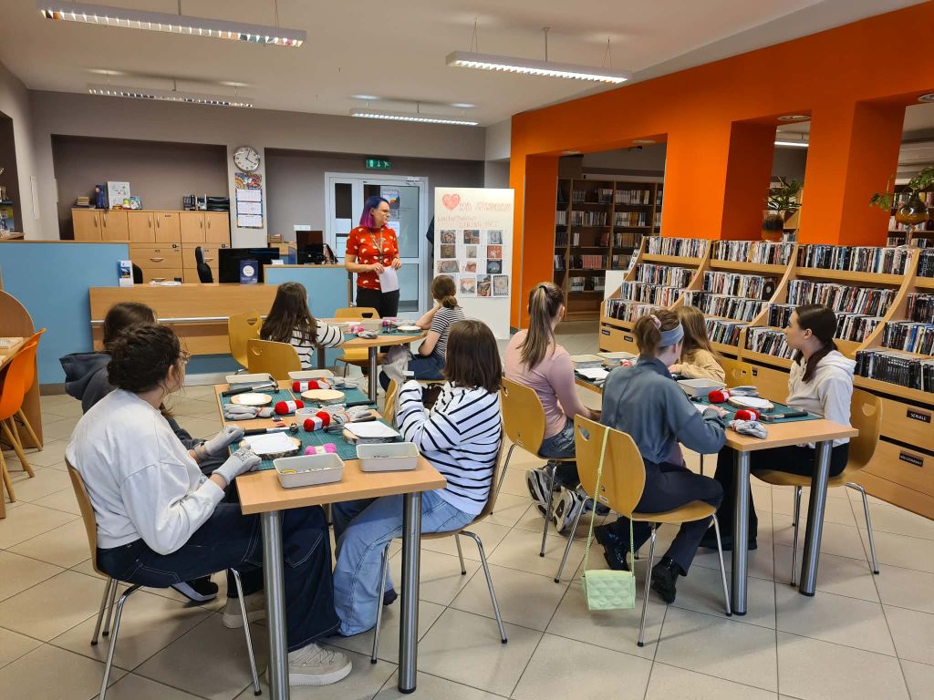 Grupa młodzieży siedzi przy stolikach w nowoczesnej, jasnej bibliotece z pomarańczową ścianą. Przed nimi leżą materiały do rękodzieła. Na środku stoi instruktorka w czerwonej koszulce, która tłumaczy zadanie. W tle widać regały z książkami oraz tablicę z napisem Serce na sznurku - walentynkowy string art
