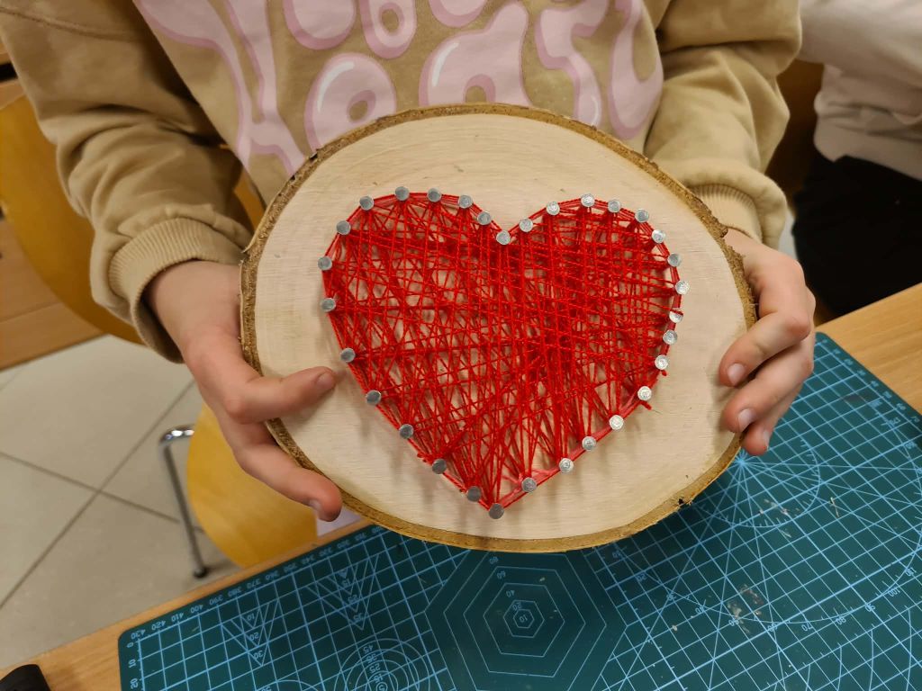 Zbliżenie na dłonie trzymające plaster drewna z korą. Na drewnie wbite są gwoździe ułożone w kształt serca, przez które gęsto przepleciony jest jaskrawoczerwony sznurek. Pod dłońmi widoczna jest zielona mata do cięcia z podziałką.