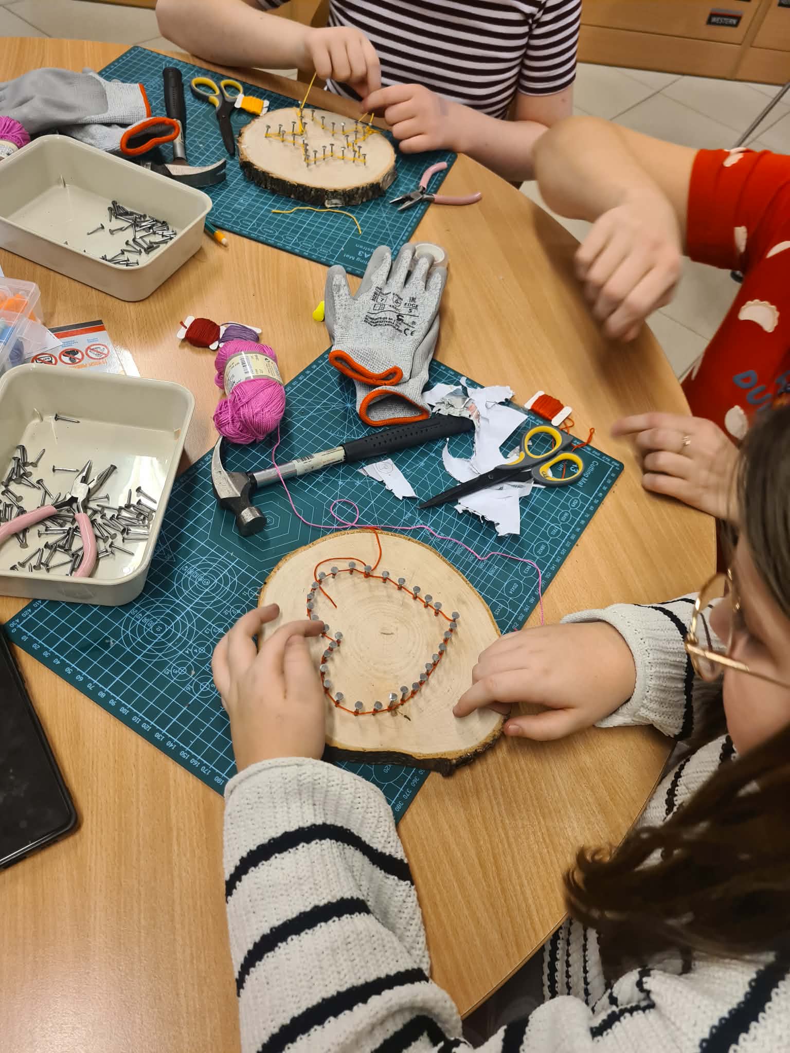Widok z góry na stół, przy którym pracuje nastolatka. Na zielonej macie leży plaster drewna z wbitymi gwoździami tworzącymi kontur serca. Wokół rozsypane są narzędzia: młotek, nożyczki, szczypce, rękawica ochronna oraz motki różowej i czerwonej włóczki.