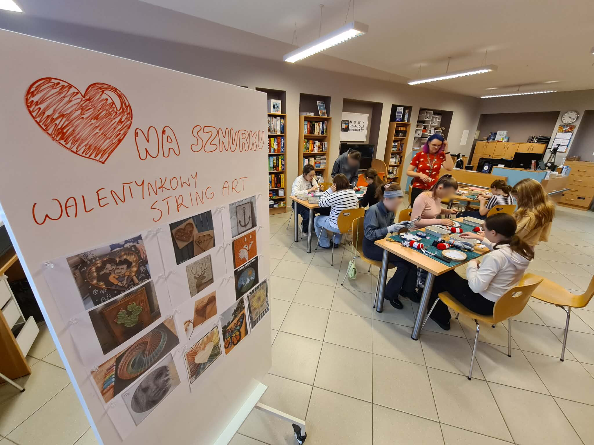 Szerokie ujęcie sali bibliotecznej. Na pierwszym planie biała tablica z dużym, narysowanym sercem i napisem: „Serce na sznurku – walentynkowy string art”. Do tablicy przypięte są zdjęcia inspiracji. W głębi widać młodzież siedzącą przy stolikach i regały z grami planszowymi.