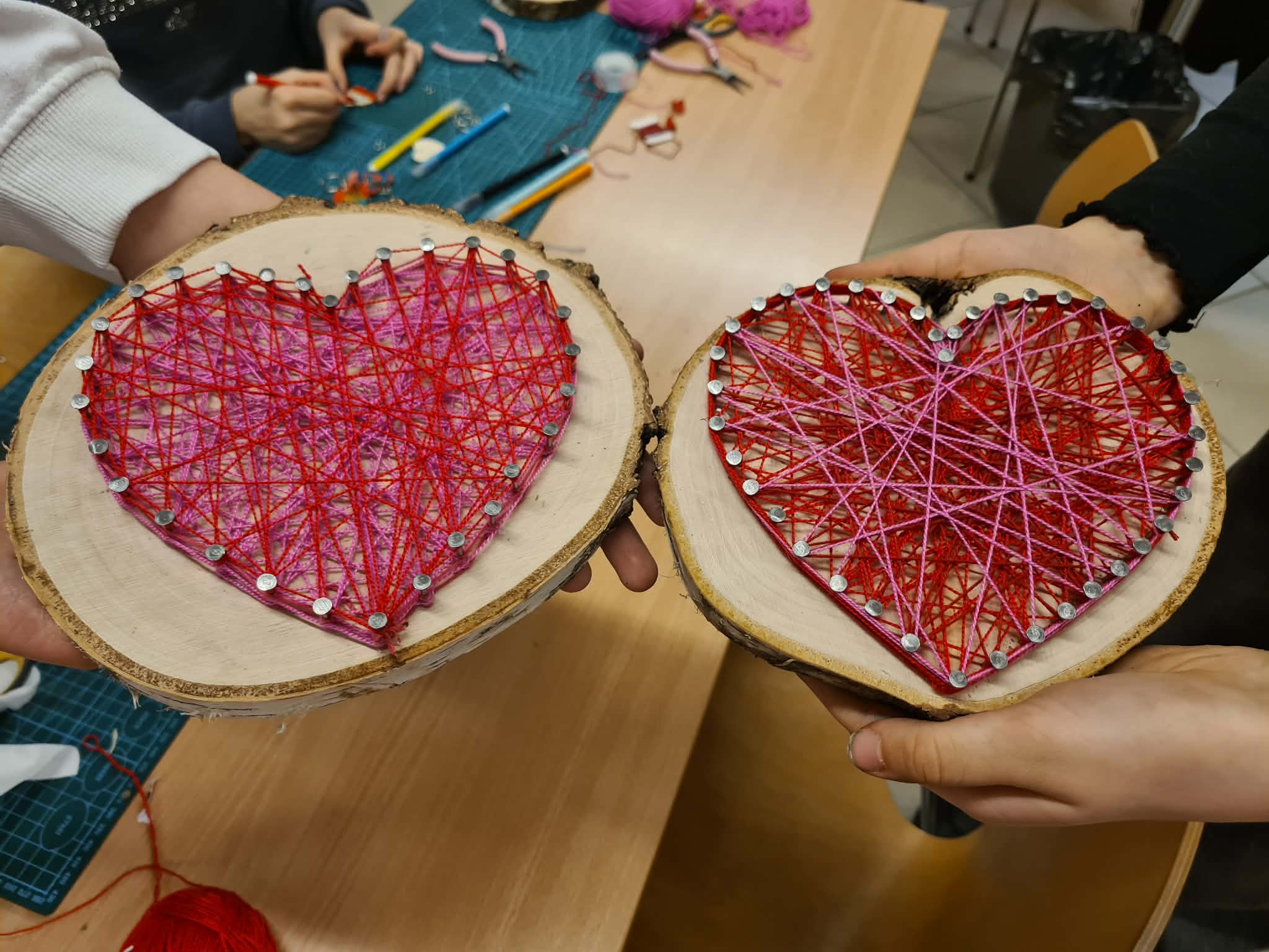 Dwie osoby trzymają obok siebie swoje gotowe prace na plastrach drewna. Serce po lewej stronie jest wykonane z jaśniejszej, różowo-czerwonej włóczki, a serce po prawej ma ciemniejsze, intensywnie czerwone wypełnienie z różowym akcentem na krawędzi.