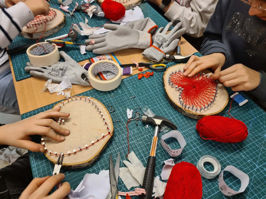 Widok z góry na stół warsztatowy. Dwie osoby przygotowują bazy do swoich prac: jedna przykleja papierowy szablon serca na plastrze drewna, a druga wbija gwoździe wzdłuż krawędzi swojego plastra za pomocą młotka. Na stole leżą motki czerwonej włóczki i rękawice ochronne.