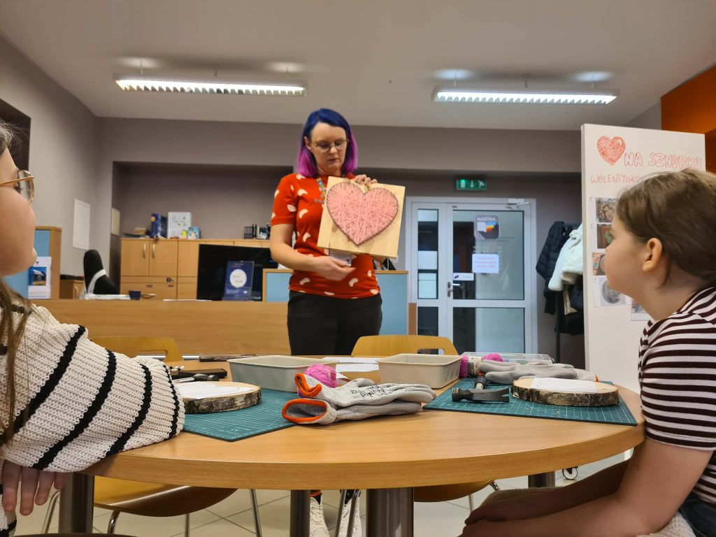 Instruktorka z niebiesko-fioletowymi włosami stoi przed uczestnikami i prezentuje gotowy wzór serca wykonany techniką string-art na kwadratowej desce. Uczestnicy siedzący przy okrągłym stole obserwują pokaz. Na stole leżą przygotowane plastry drewna i szare rękawice robocze.