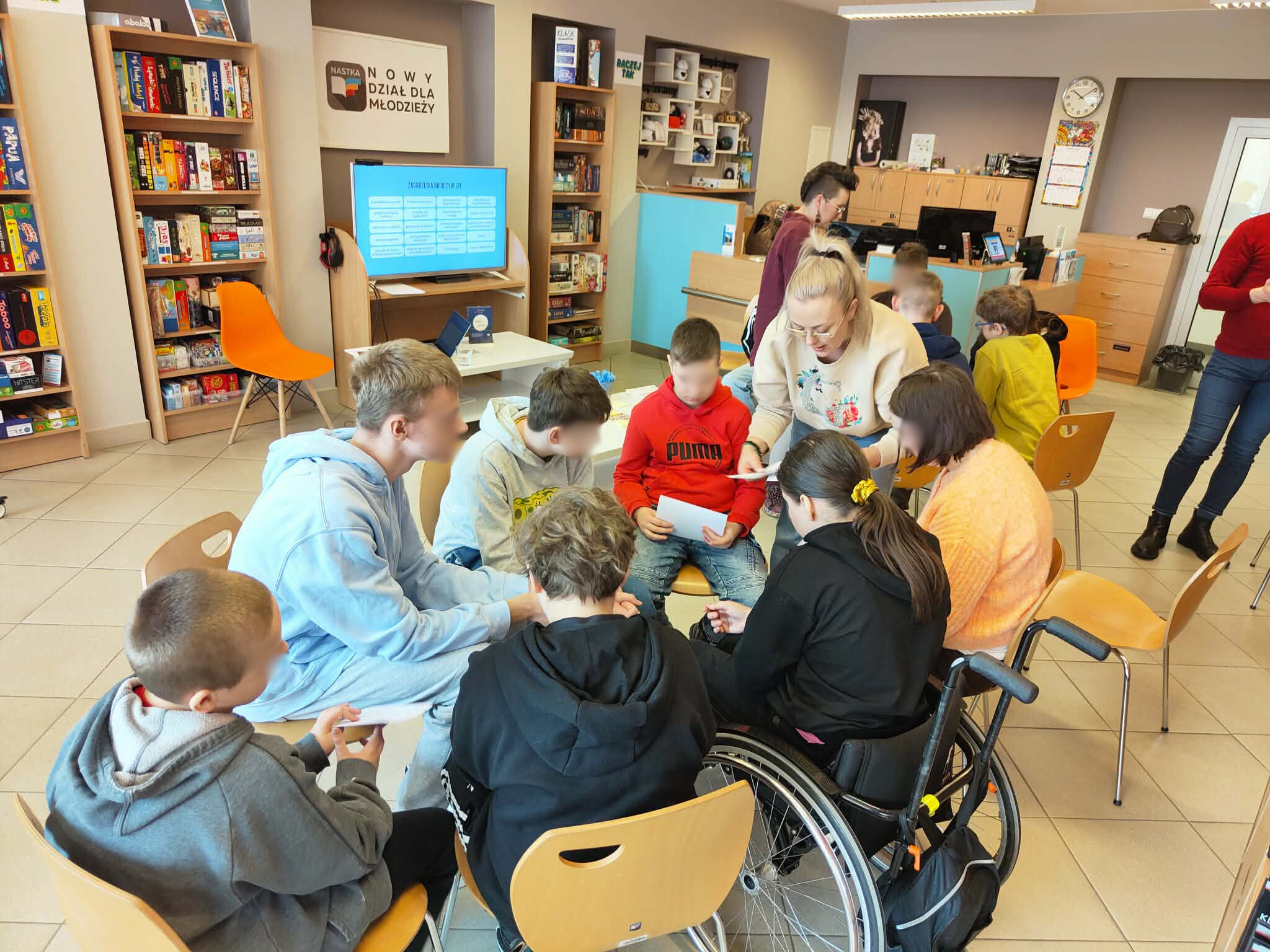 Uczestnicy warsztatów pracują w grupie, jedna z osób korzysta z wózka inwalidzkiego, prowadząca omawia zadanie.