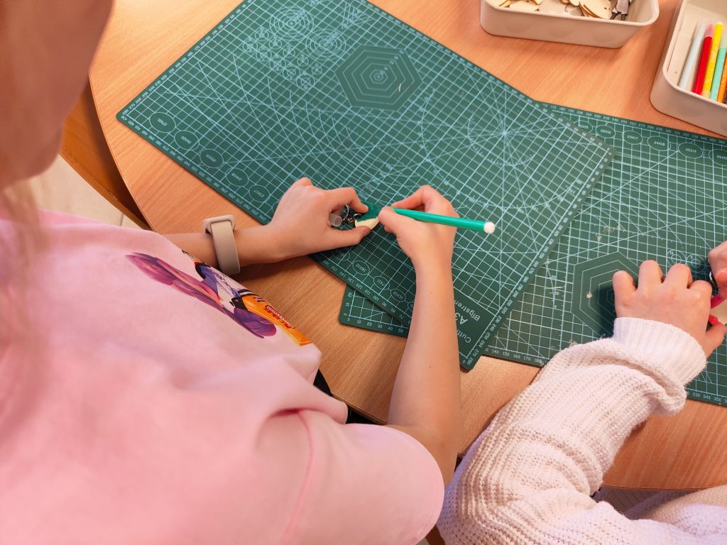 Zbliżenie na dłonie osoby w różowej koszulce, która ozdabia małe drewniane serce zielonym mazakiem. Osoba ma na nadgarstku jasny zegarek typu smartwatch. W tle widoczny fragment zielonej maty i biały pojemnik z mazakami.