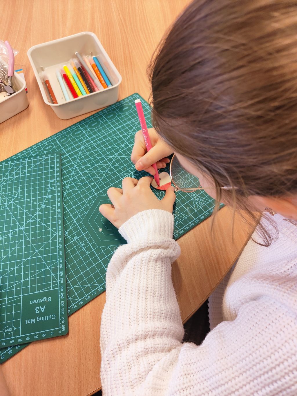 Zbliżenie z perspektywy ramienia na osobę w białym swetrze, która precyzyjnie koloruje dolną część drewnianego serduszka różowym flamastrem. Praca odbywa się na profesjonalnej zielonej macie z podziałką.