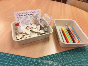 Zdjęcie przedstawia zestaw do samodzielnego tworzenia breloków w kształcie serc, umieszczony na drewnianym blacie obok zielonej maty modelarskiej. W dwóch jasnych pojemnikach znajdują się: drewniane bazy w kształcie serc, metalowe kółka z łańcuszkami, różowe szczypce jubilerskie oraz kolorowe mazaki. W jednym z pojemników stoi kartka z napisem „SERCE DO WZIĘCIA” i instrukcją wykonania.