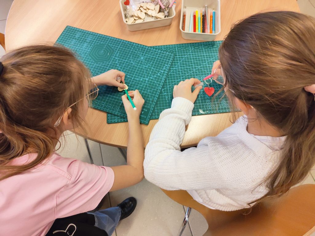Widok z góry na dwie osoby siedzące przy drewnianym stole. Na zielonych matach modelarskich każda z nich koloruje drewniane serduszko – jedna używa zielonego, a druga różowego mazaka. W tle widać pojemniki z materiałami i instrukcję „Serce do wzięcia”.