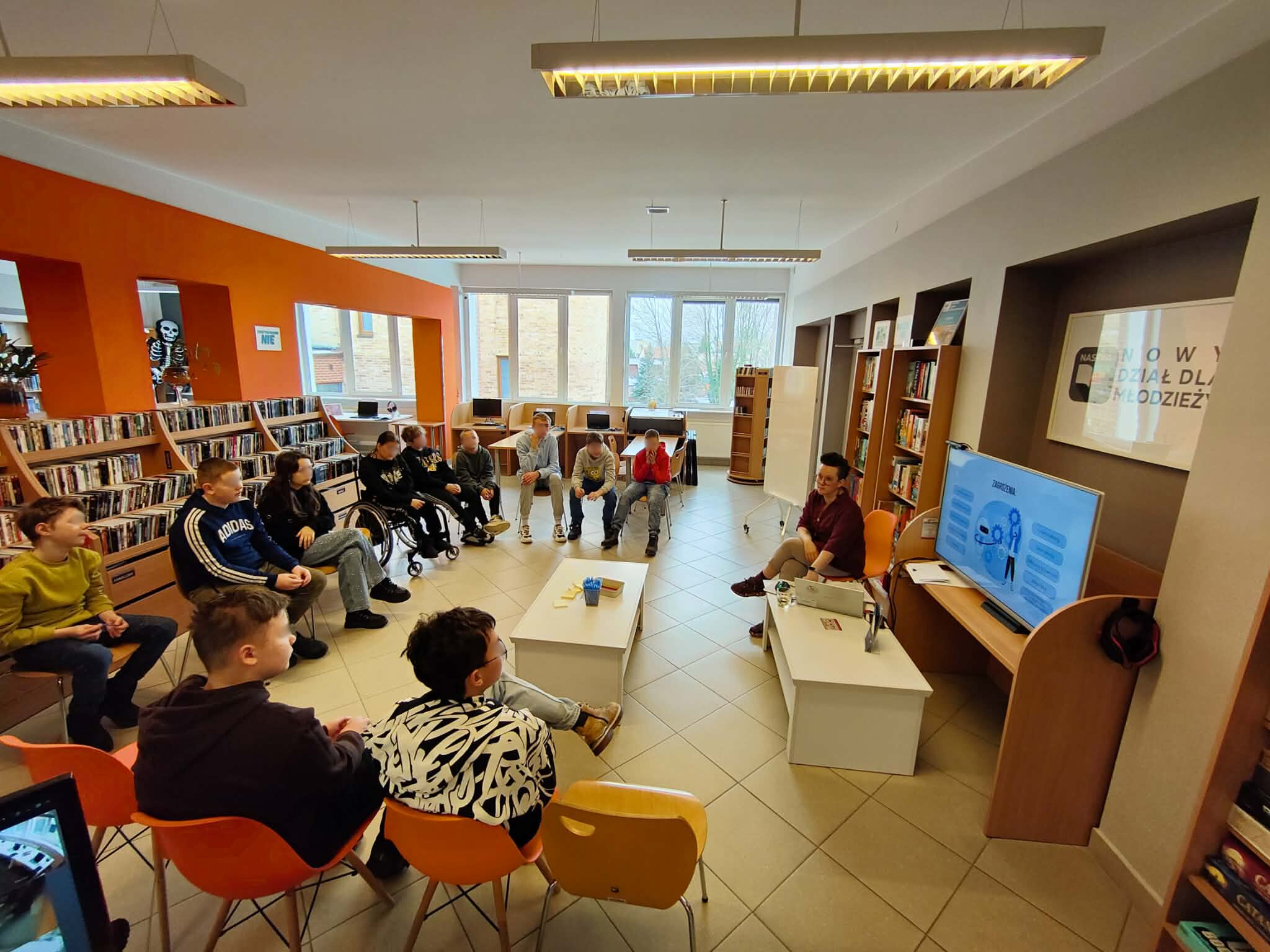 Grupa młodzieży siedzi w kręgu i słucha prowadzącej prezentującej treści na ekranie.