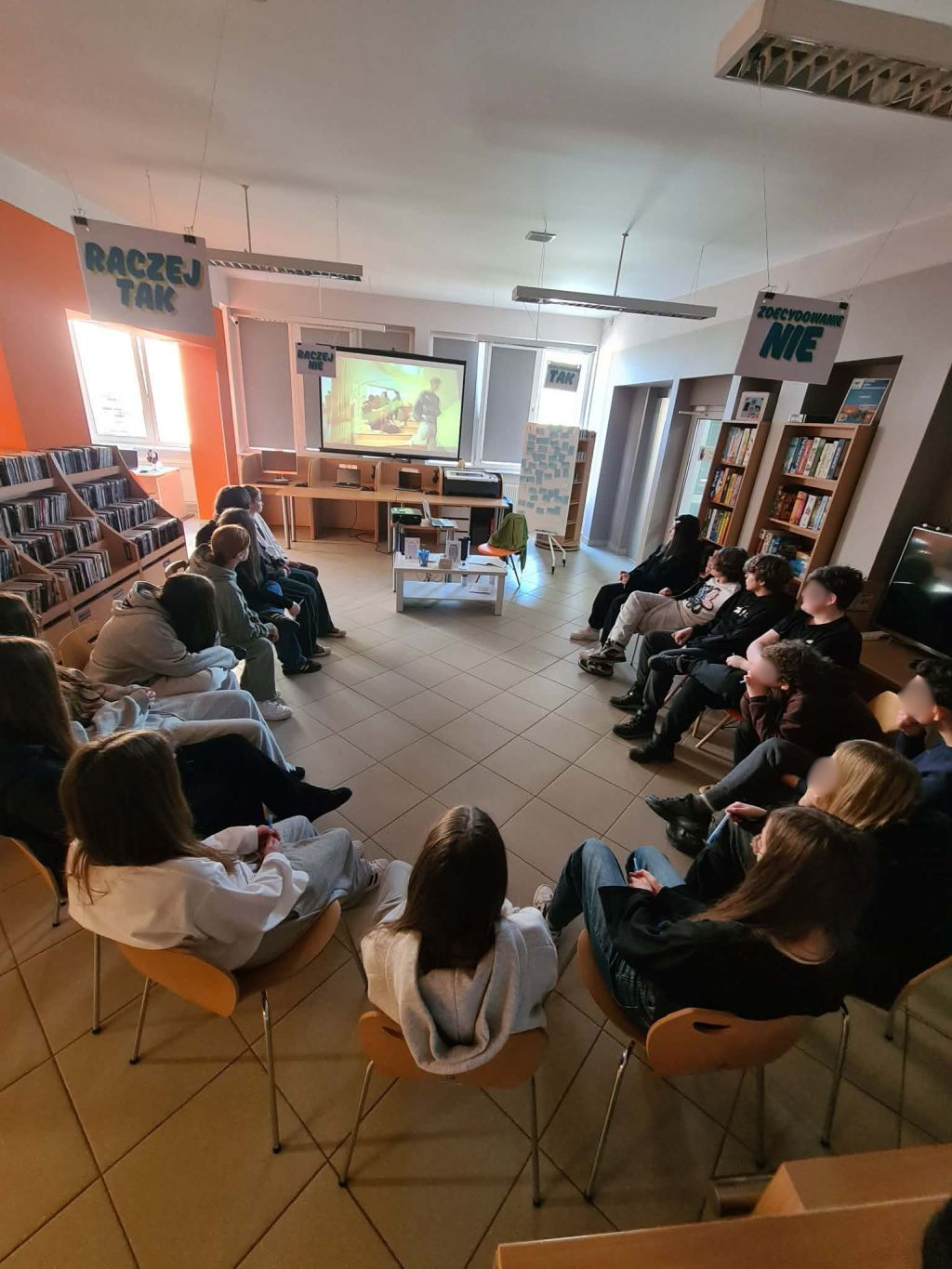 Grupa młodzieży siedzi w kręgu na krzesłach w nowoczesnej bibliotece. Nad nimi wiszą tabliczki z napisami „RACZEJ TAK” oraz „ZDECYDOWANIE NIE”. W tle widać ekran projekcyjny z wyświetlonym filmem oraz regały z książkami i płytami.