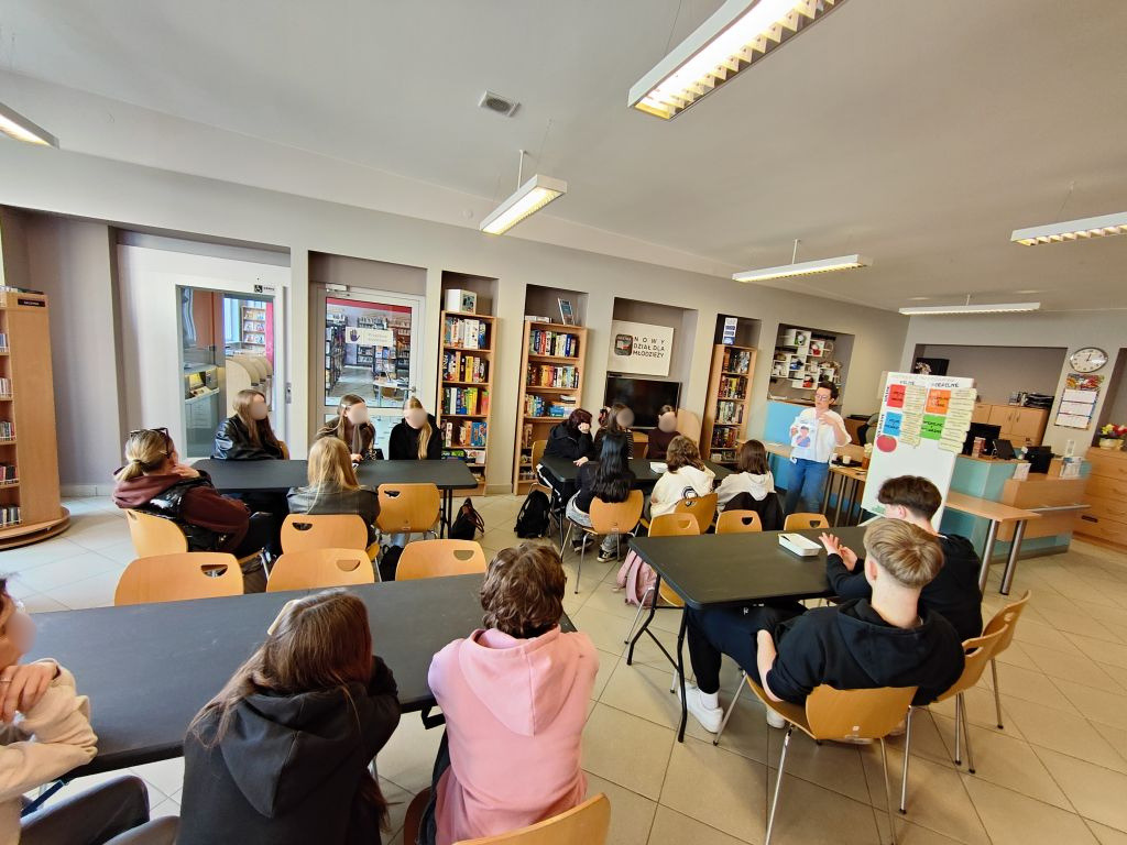 Szeroki kadr sali warsztatowej; młodzież siedzi w grupach przy stolikach, a prowadząca prezentuje materiały edukacyjne.