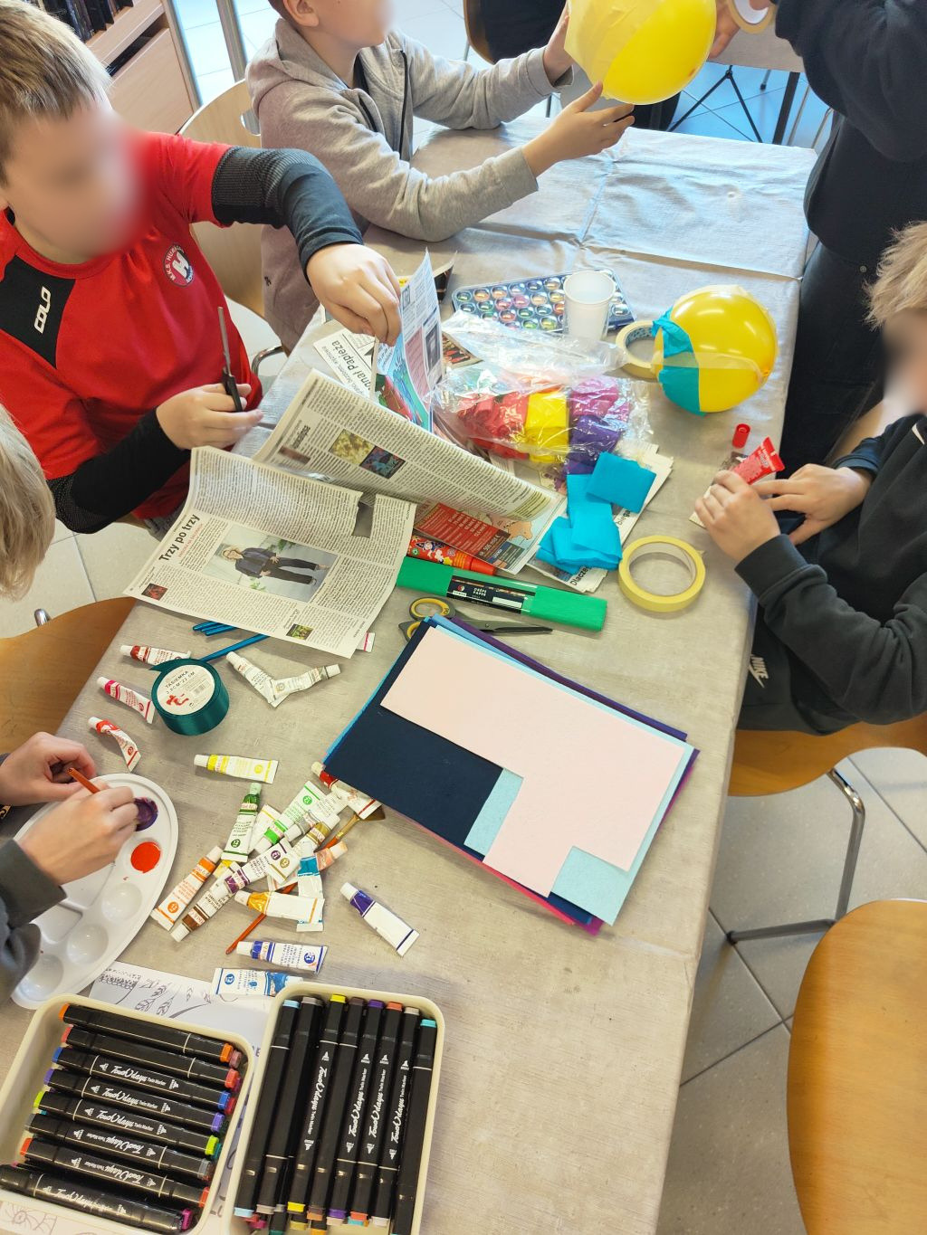 Zbliżenie na stół warsztatowy zasypany materiałami plastycznymi: farbami w tubkach, markerami, kolorową bibułą, gazetami i żółtymi balonami.