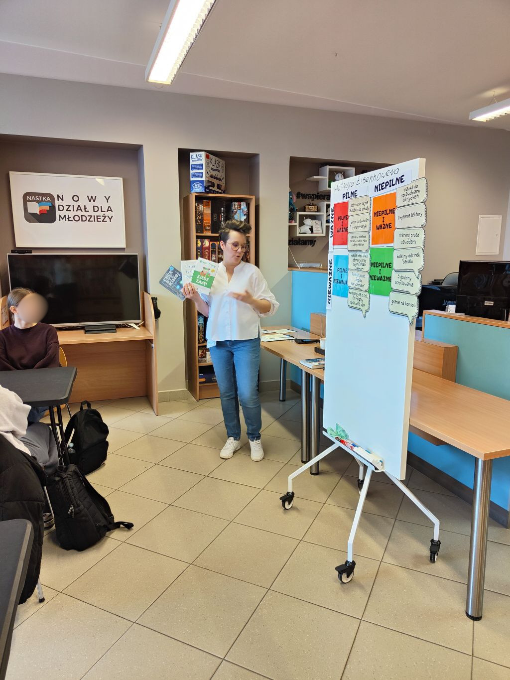 Kobieta prowadząca warsztaty trzyma w rękach książki, stojąc obok tablicy z napisem Matryca Eisenhowera i kolorowymi polami zadań.