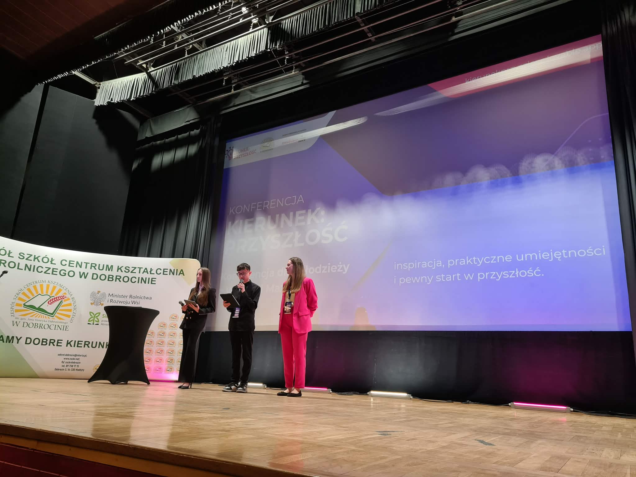 Trzy osoby stoją na scenie i prowadzą prezentację podczas konferencji dla młodzieży.