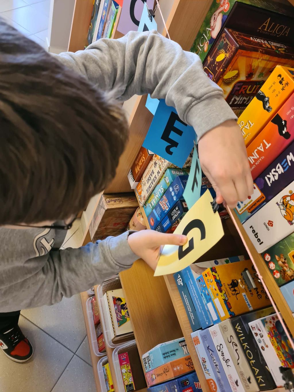 Zbliżenie na ręce dziecka przy regale z grami planszowymi, przypinającego klamerką żółtą kartkę z literą 