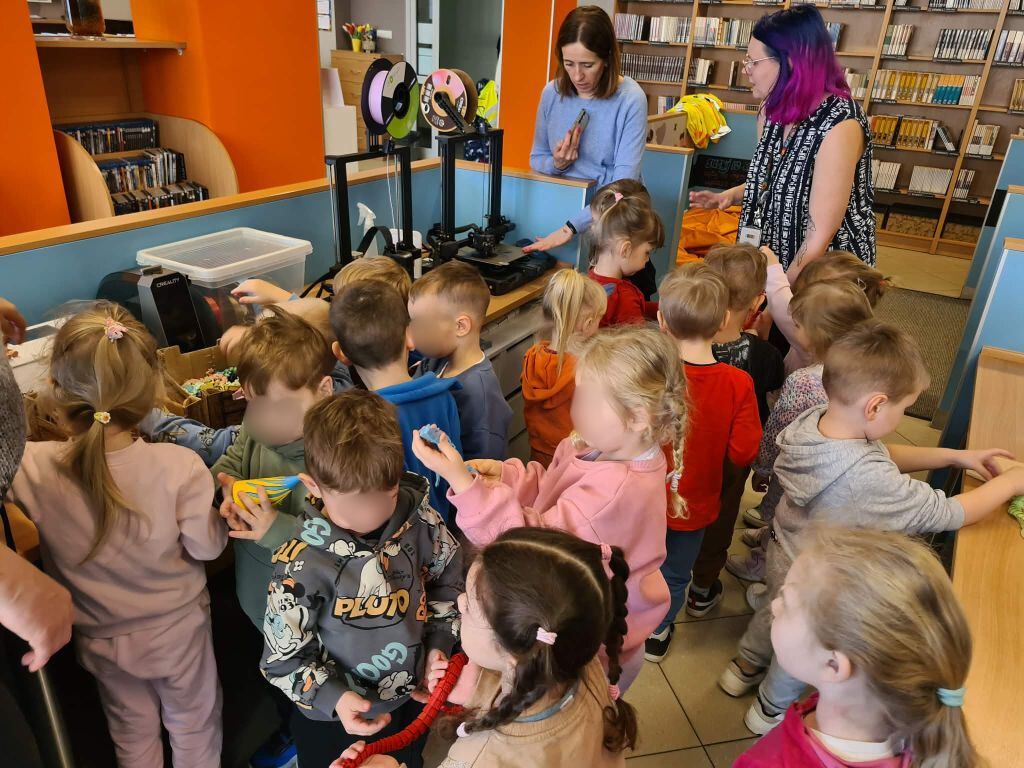 Grupa dzieci stoi stłoczona przy niebieskiej ladzie, na której umieszczone są dwie czarne drukarki 3D z założonymi szpulami kolorowego filamentu. Obok urządzeń stoi kobieta w niebieskim swetrze oraz druga z fioletowymi włosami i tatuażami na przedramionach.