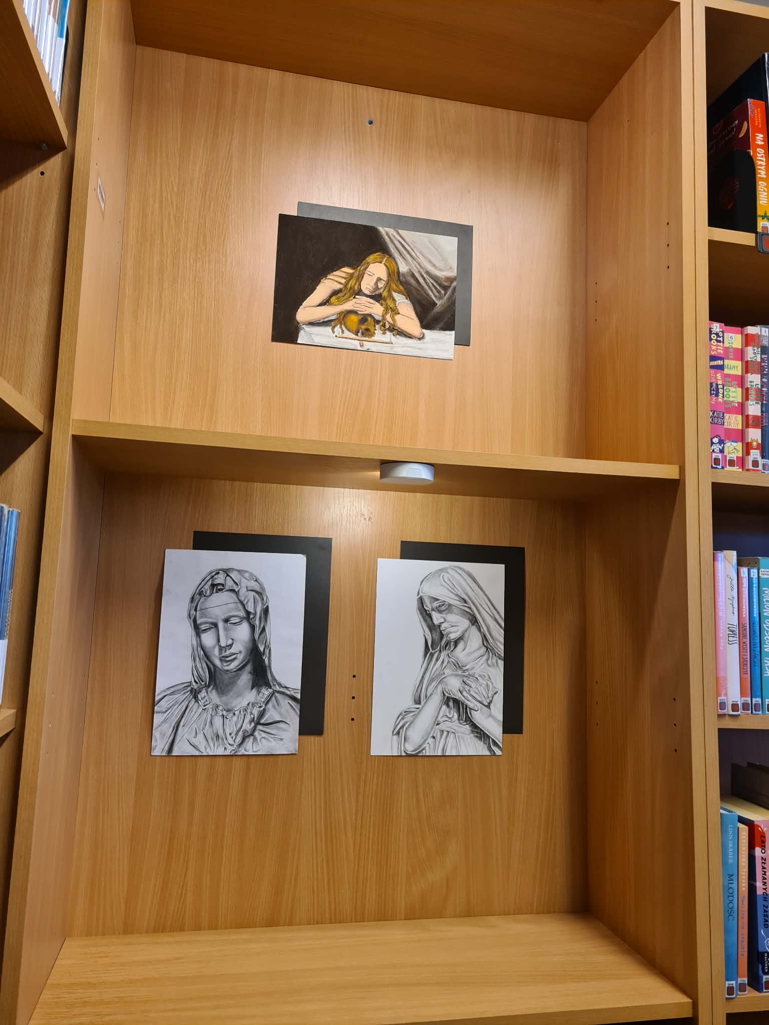 Prace plastyczne przedstawiające portrety i postacie kobiece zaprezentowane na półce bibliotecznej.