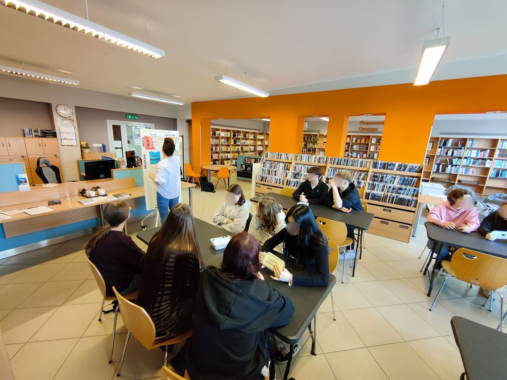 Widok z tyłu na grupę młodzieży przy stolikach w bibliotece. W głębi widać pomarańczową ścianę z regałami pełnymi książek i filmów.