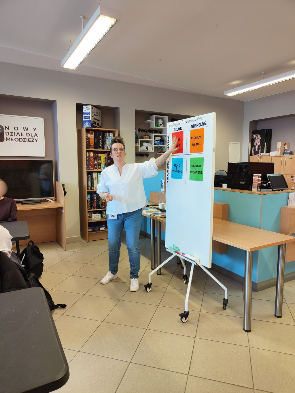 Kobieta w okularach i białej koszuli wskazuje na kolorową matrycę Eisenhowera przyczepioną do tablicy flipchart.