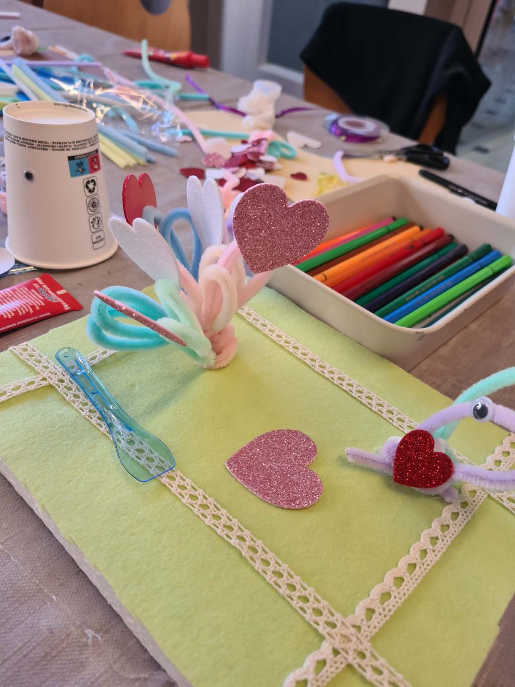 Zbliżenie na zieloną podkładkę z filcu ozdobioną koronką, na której znajdują się brokatowe serca i dekoracje z kolorowych drucików kreatywnych. W tle widać kubek, mazaki i inne przybory plastyczne.
