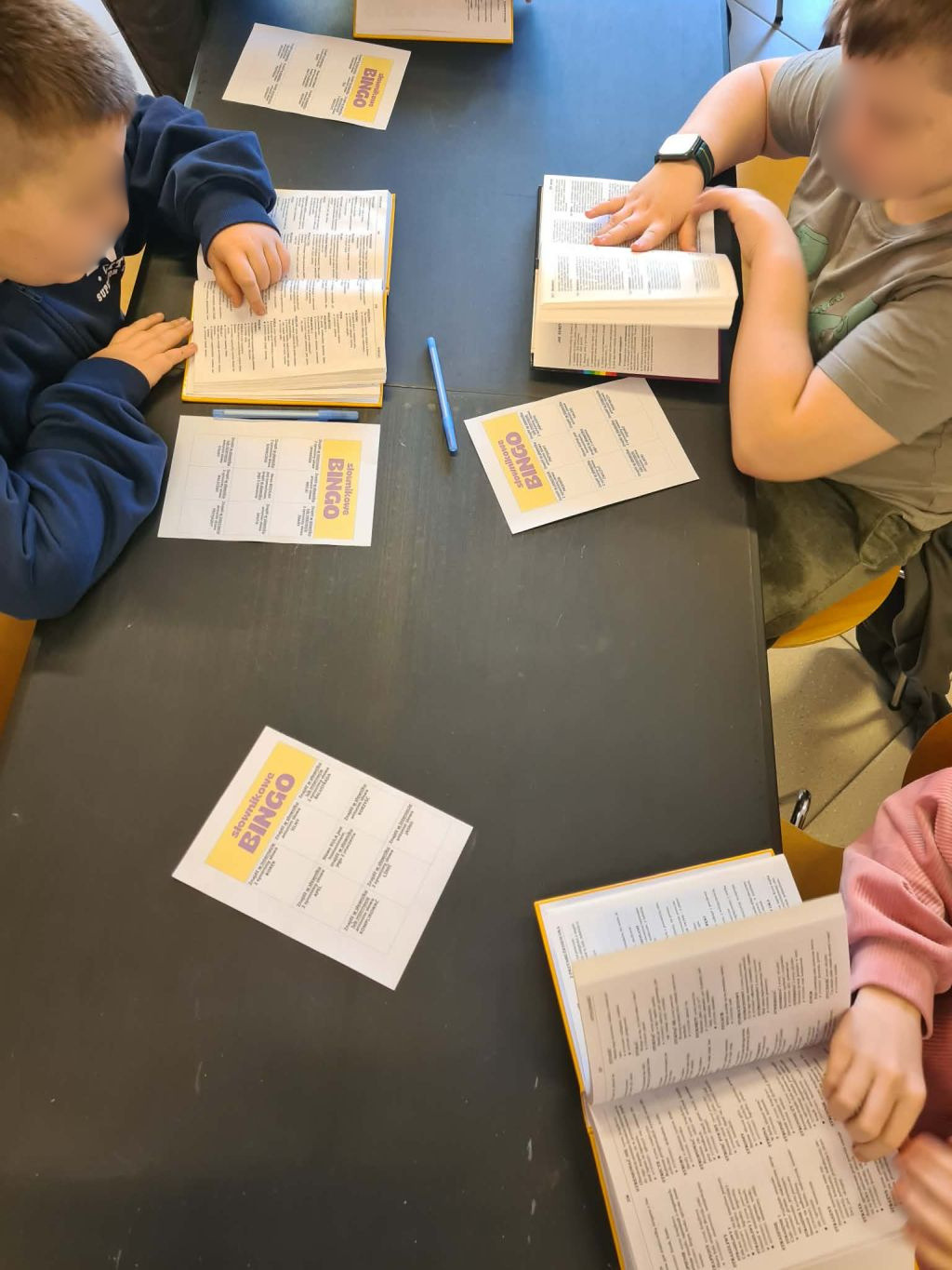 Widok z góry na troje dzieci siedzących przy czarnym stole, z których dwoje czyta otwarte książki, a jedno trzyma dłoń na swojej książce, z kartkami BINGO przed nimi.