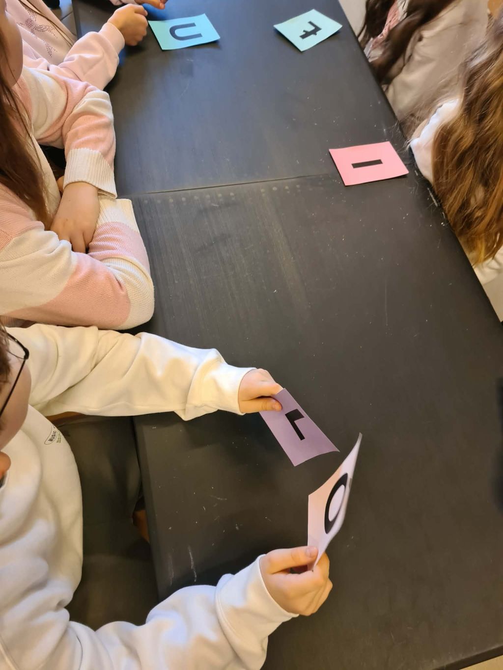 Dzieci przy czarnym blacie trzymają kolorowe kartki z pojedynczymi, dużymi literami.