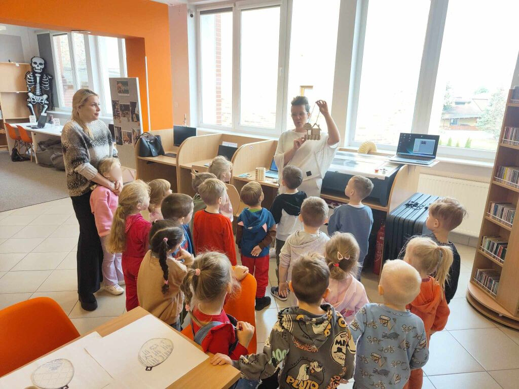 Dzieci stoją w półkolu przed kobietą w białej bluzce, która trzyma w górze ażurową, drewnianą ozdobę w kształcie koszyczka. Spotkanie odbywa się w jasnym pomieszczeniu z dużymi oknami, przy których znajdują się stanowiska komputerowe.