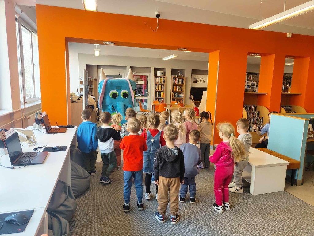 Grupa dzieci stoi tyłem do obiektywu na szarej wykładzinie w nowoczesnej sali bibliotecznej z jaskrawopomarańczowymi ścianami. Dzieci patrzą w stronę niebieskiej maskotki słonia z dużymi oczami, która stoi w przejściu do dalszej części biblioteki. Po lewej stronie widoczny jest biały blat ze stanowiskami komputerowymi i laptopami, a w głębi regały z książkami.