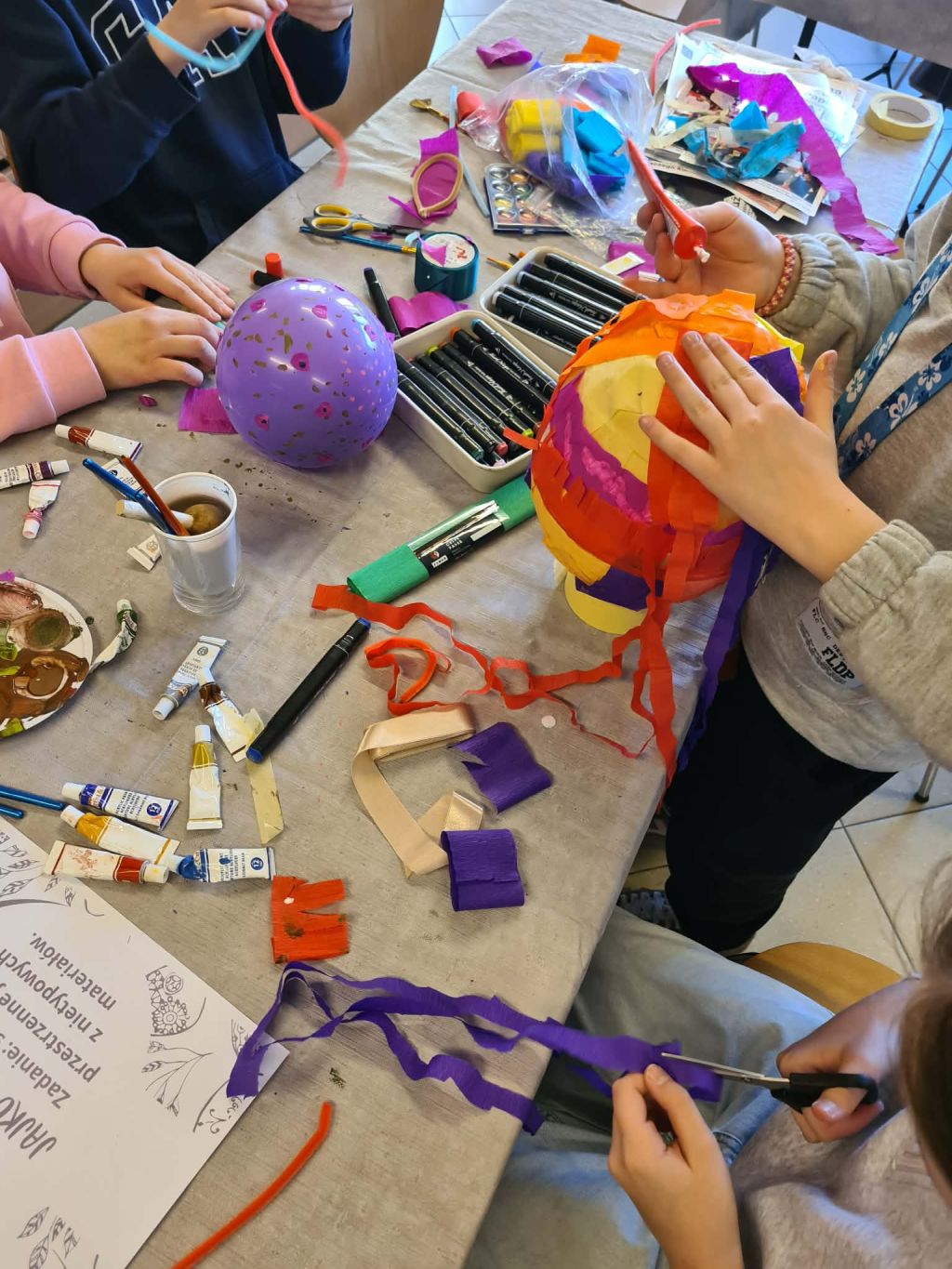 Proces tworzenia formy przestrzennej; dziecko przykleja paski pomarańczowej bibuły do kuli, tworząc kolorową strukturę obok innych przyborów plastycznych.