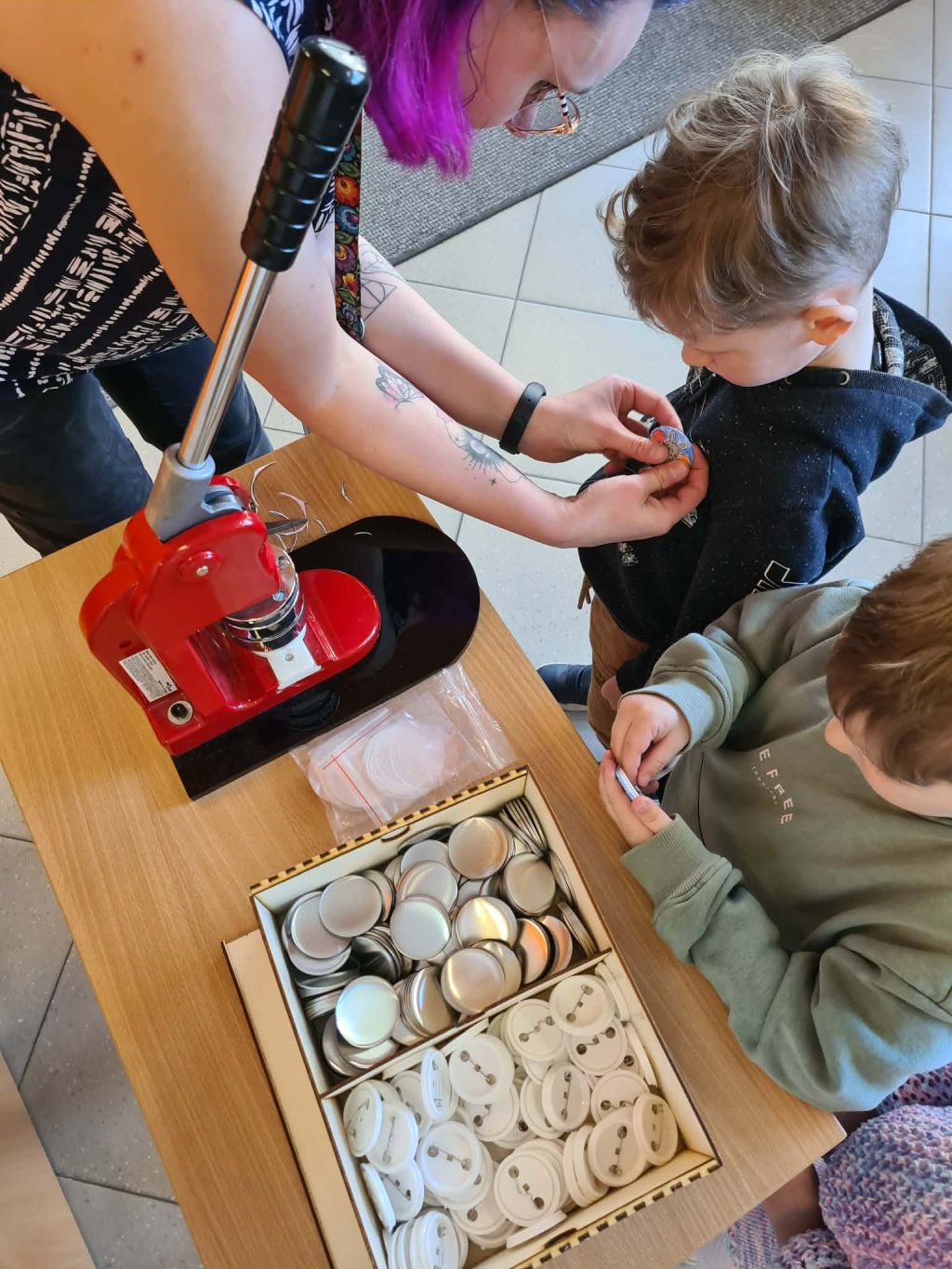 Widok z góry na stół, przy którym kobieta z fioletowymi włosami przypina dziecku gotowy znaczek. Obok na stole stoi czerwona prasa do robienia przypinek oraz drewniane pudełko z metalowymi elementami i agrafkami.