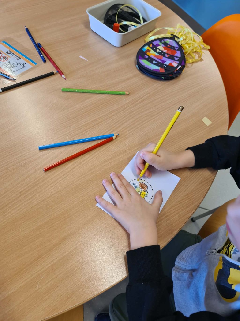 Dziecko siedzi przy okrągłym stole i koloruje żółtą kredką rysunek zająca z pisanką. Na stole leżą luźno rozrzucone kredki w różnych kolorach oraz gotowa, wyklejona kolorową bibułą ozdoba z doczepionymi żółtymi wstążkami.
