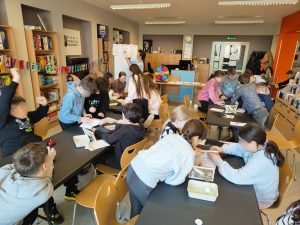 Ujęcie z góry na pełną salę biblioteczną, gdzie grupy dzieci siedzą przy czarnych stołach, a prelegentka stoi w głębi przy białej tablicy.