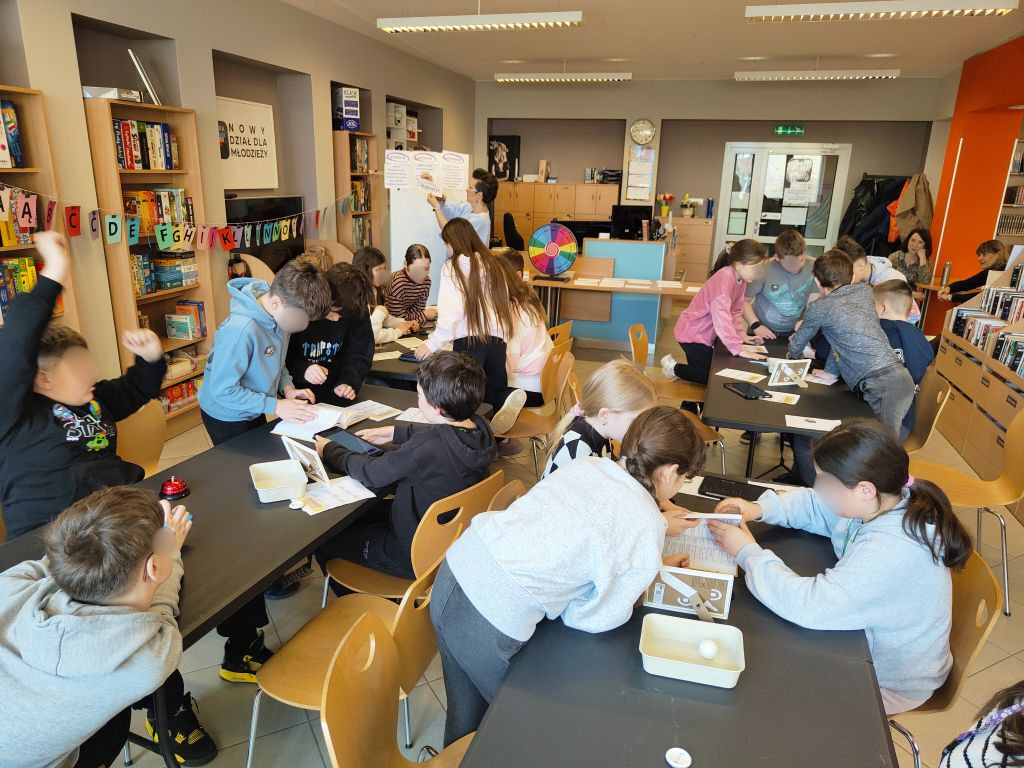 Ujęcie z góry na pełną salę biblioteczną, gdzie grupy dzieci siedzą przy czarnych stołach, a prelegentka stoi w głębi przy białej tablicy.
