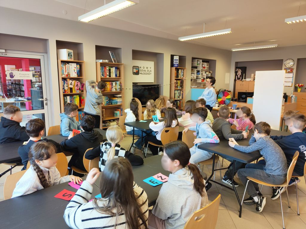 Szeroki widok na salę biblioteczną z grupą dzieci siedzących przy czarnych stołach i pracujących z kolorowymi kartami, prelegentka stoi w głębi, na regałach widać girlandę z liter.