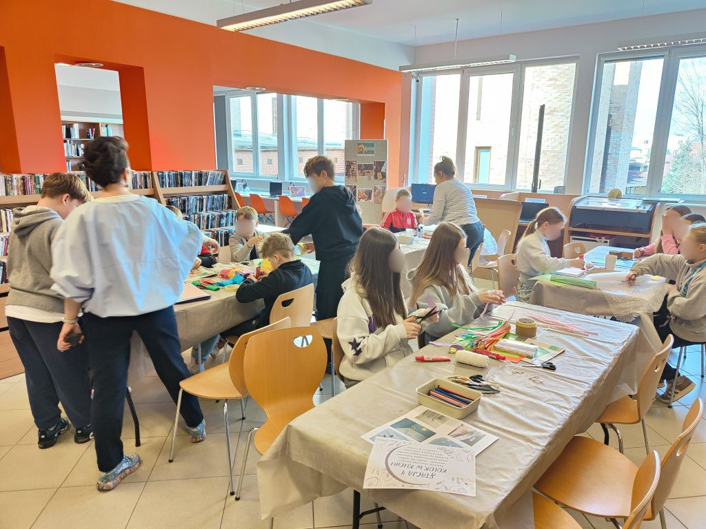Szeroki kadr sali bibliotecznej, w której dzieci przy kilku stołach wycinają, kleją i malują, tworząc prace plastyczne.