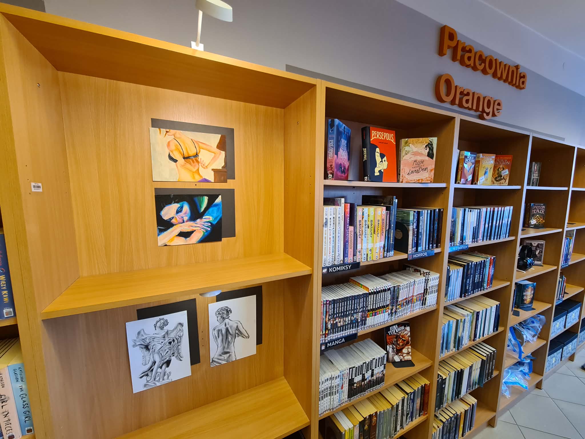 Fragment wystawy prac plastycznych na regale w dziale młodzieżowym NASTKA, obok półek z książkami i napisu „Pracownia Orange”.