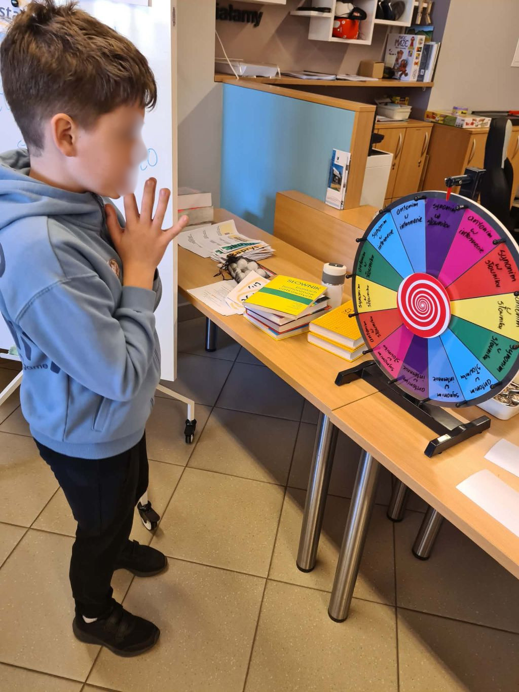 Chłopiec w błękitnej bluzie stoi z rękami złożonymi razem, patrząc na kolorowe koło fortuny z napisami stojące na stole w bibliotece.