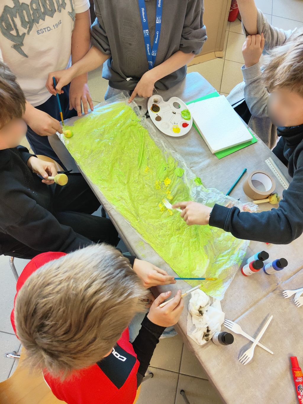 Widok z góry na grupę dzieci wspólnie malujących pędzlami i gąbkami duży arkusz folii rozłożony na stole, używając jasnozielonej farby.